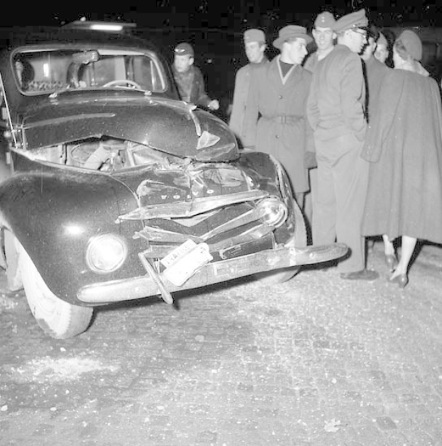 Schwarz-weißes Bild eines schwer beschädigten Autos am Straßenrand, umgeben von einer Gruppe von Menschen.