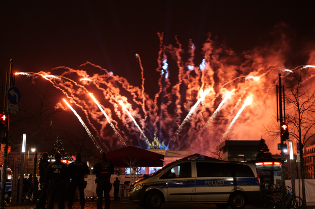 Polizeiauto vor Menschenmenge während der Neujahrsfeier in Berlin geparkt, mit farbigen Feuerwerken am Himmel und Stadtlichtern im Hintergrund.