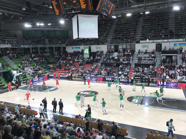 Eine Gruppe von Menschen, die Basketball in einer vollen Arena mit Zuschauern in den Rängen spielt, beleuchtet von Stadionlampen und umgeben von Anzeigetafeln.