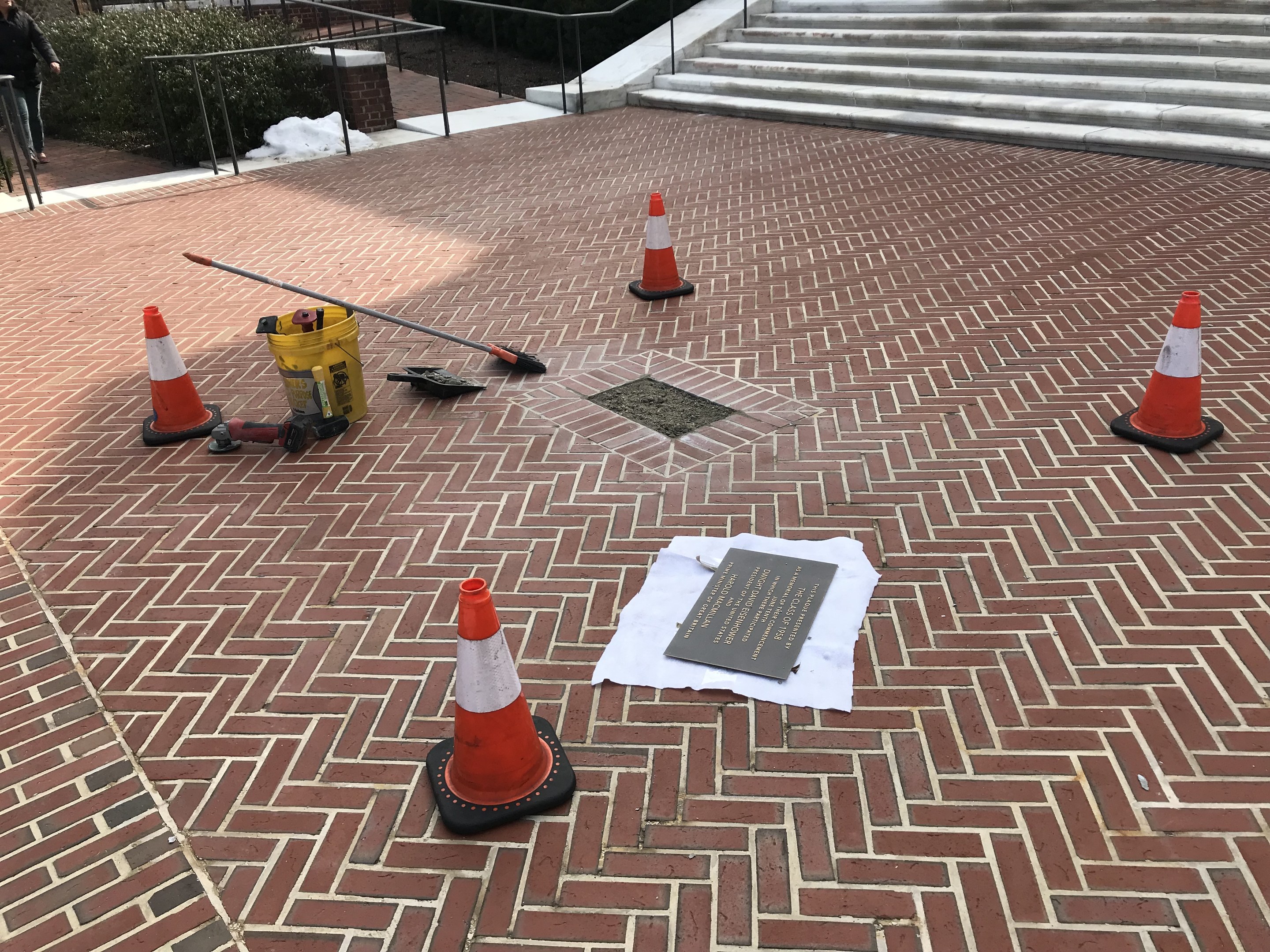 Backsteinweg mit Verkehrskegeln, einem Schild und einer Person auf der linken Seite, umgeben von Pflanzen, Geländern und Treppen, mit Bäumen und einem Gebäude im Hintergrund.