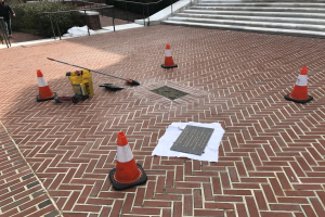 Backsteinweg mit Verkehrskegeln, einem Schild und einer Person auf der linken Seite, umgeben von Pflanzen, Geländern und Treppen, mit Bäumen und einem Gebäude im Hintergrund.