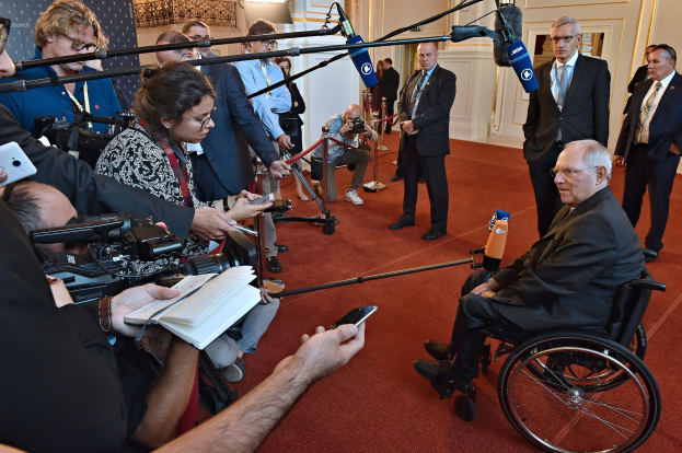 Ein Mann im Rollstuhl umgeben von Reportern mit Kameras, Mikrofonen und Handys, mit einem Banner, Scheinwerfern und einer Wand im Hintergrund.