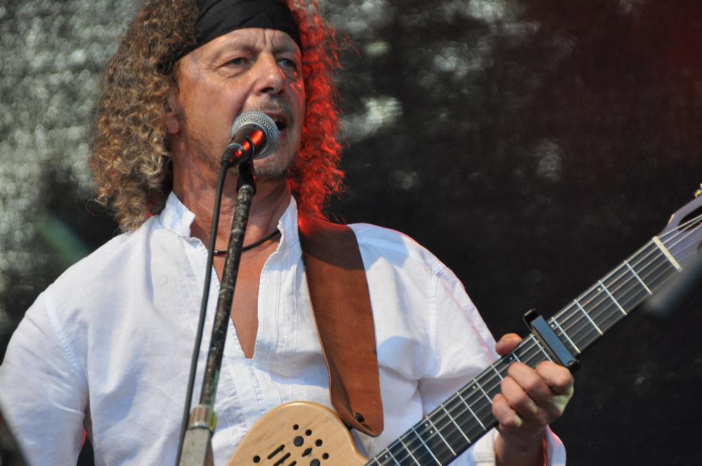 Ein Mann in einem weißen Hemd und schwarzem Stirnband singt in ein Mikrofon und hält eine Gitarre.