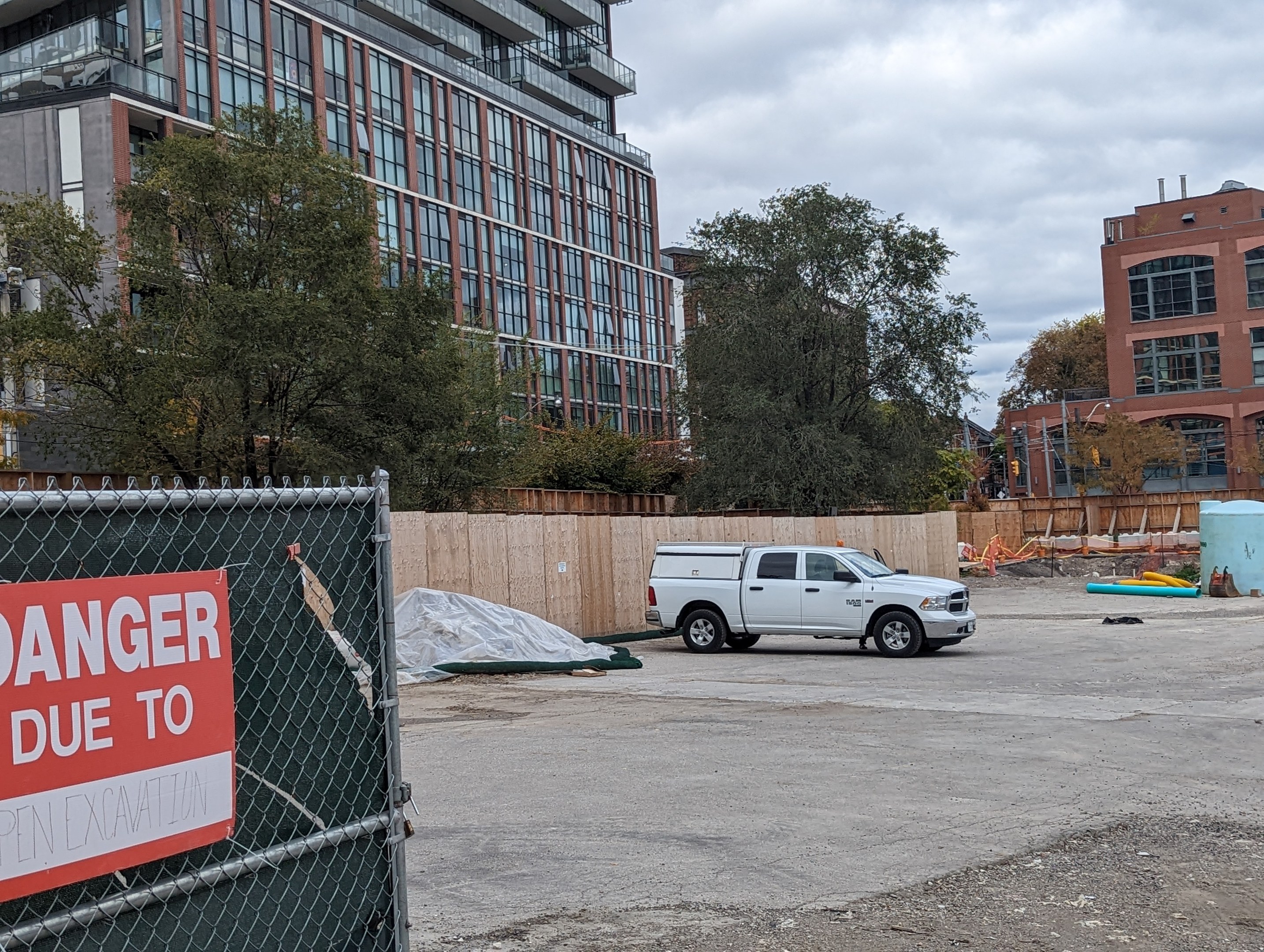 Truck parked near a building with a "Danger Due to Open Excavation" sign, trees, buildings, poles, and a clear blue sky in the background.