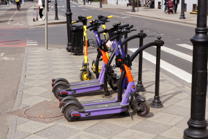 Eine Reihe von Elektro-Scootern, die auf der Straßenseite geparkt sind, mit Menschen, die auf dem Gehweg laufen und Fahrräder fahren, einem Mülleimer im Vordergrund und Gebäuden, Bäumen und Pflanzen im Hintergrund.