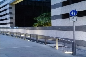 Eine Fußgängerbrücke mit einem Schild, beleuchtet von Lichtern, umgeben von Gebäuden und Bäumen, mit einem Pfahl und einer Platte und Text unten.