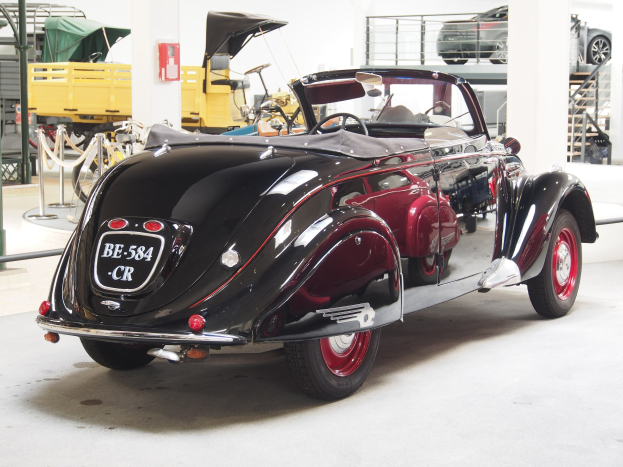 Ein schwarzer und roter Wagen in einer Garage mit anderen Fahrzeugen, Pfosten, Ketten und Gegenständen, mit einem Feuerlöscher an der Wand im Hintergrund.