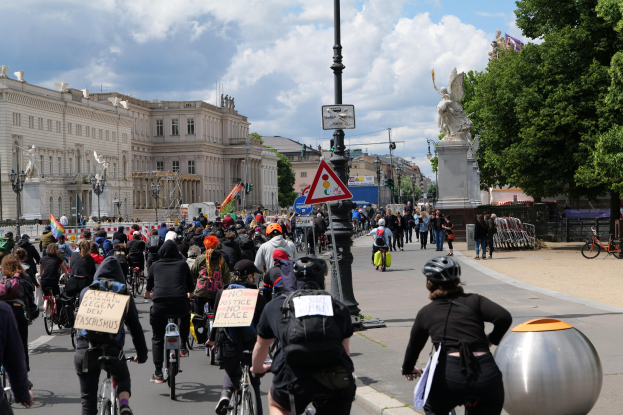 Eine große Gruppe von Radfahrern fährt eine von hohen Gebäuden gesäumte Stadtstraße entlang, einige tragen Helme und haben Taschen dabei, andere halten Schilder, Stangen, Schilder, eine Statue, Bäume und einen bewölkten Himmel im Hintergrund.