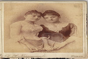 Zwei junge Frauen in traditioneller russischer Kleidung posen auf einem alten Foto, eine in einem weißen Kleid und die andere in einem blauen Kleid, vor einem hellblauen Hintergrund mit Text unten.