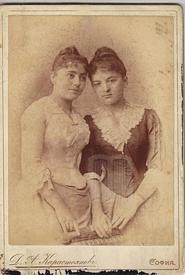 Zwei junge Frauen in traditioneller russischer Kleidung posen auf einem alten Foto, eine in einem weißen Kleid und die andere in einem blauen Kleid, vor einem hellblauen Hintergrund mit Text unten.