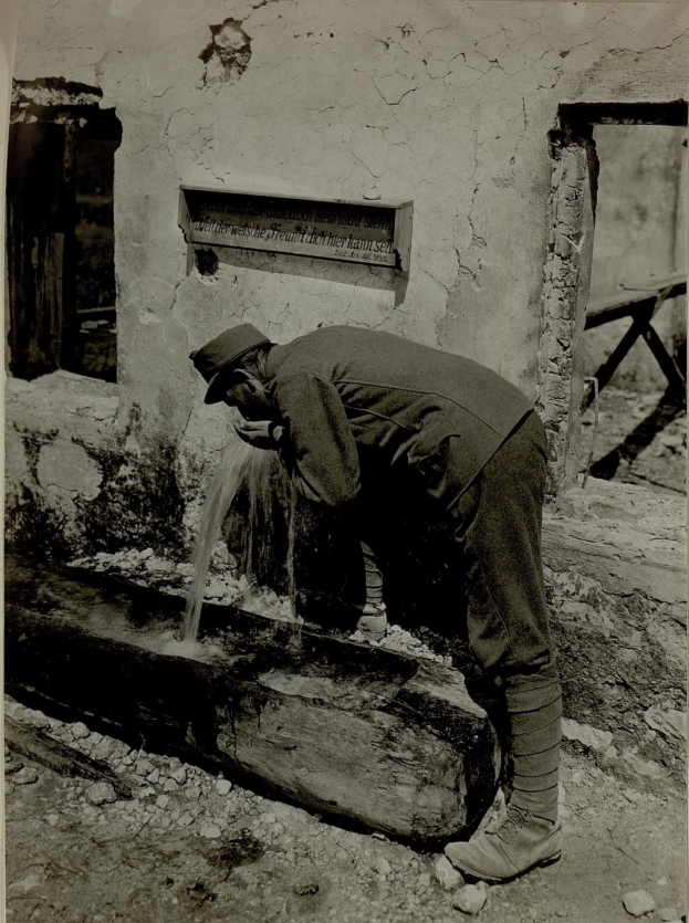Ein altes Schwarz-Weiß-Foto eines Mannes in Hut und Schuhen, der Wasser aus einem Brunnen trinkt, mit einer Wand, einem Fenster und einer Tafel mit Schrift hinter ihm.