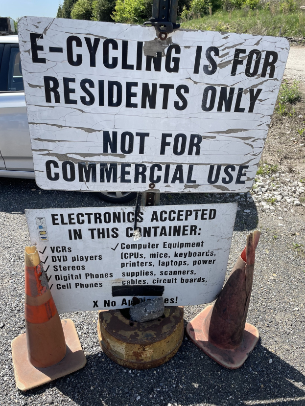 Schild mit Aufschrift "E-Bike-Fahren nur für Anwohner" neben einer Straße mit Verkehrshütchen, einem Fahrzeug, Bäumen und einem klaren blauen Himmel im Hintergrund.