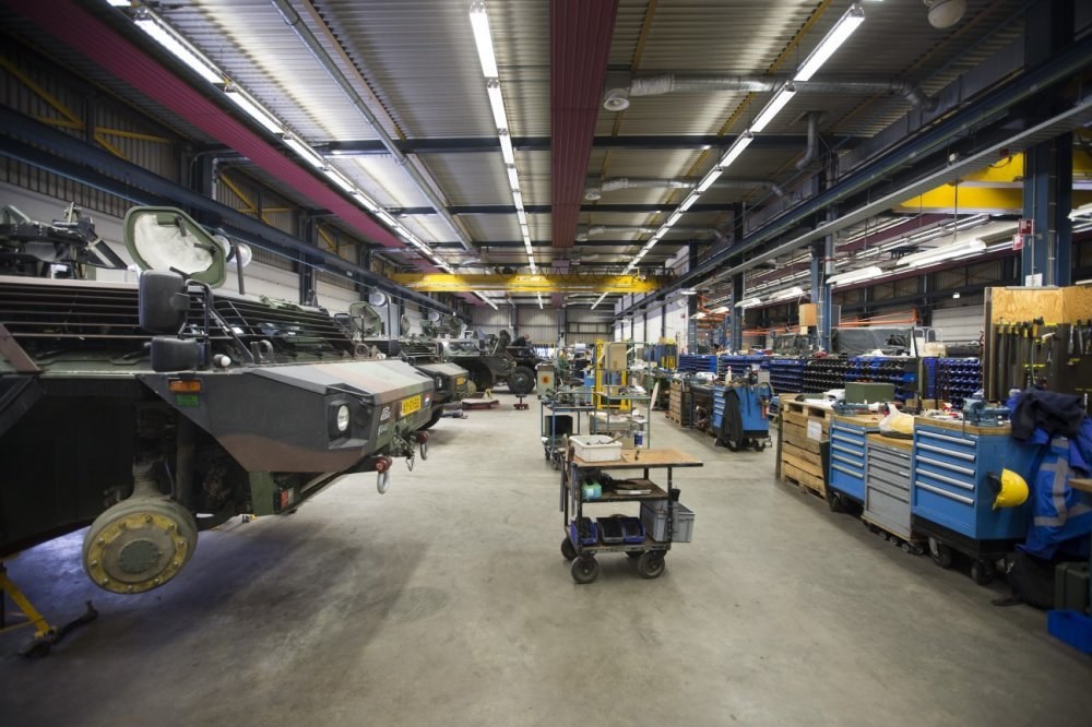 Großes Lager mit Maschinen, Fahrzeugen auf der linken Seite, Transportwagen mit Gegenständen und Holzboxen auf der rechten Seite und Deckenbeleuchtung.