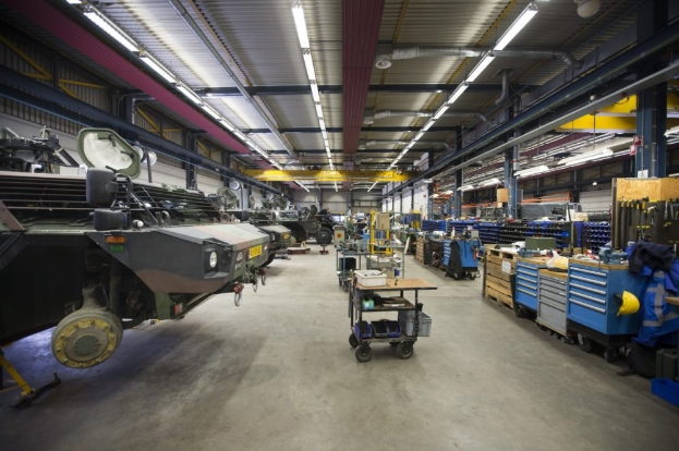 Großes Lager mit Maschinen, Fahrzeugen auf der linken Seite, Transportwagen mit Gegenständen und Holzboxen auf der rechten Seite und Deckenbeleuchtung.