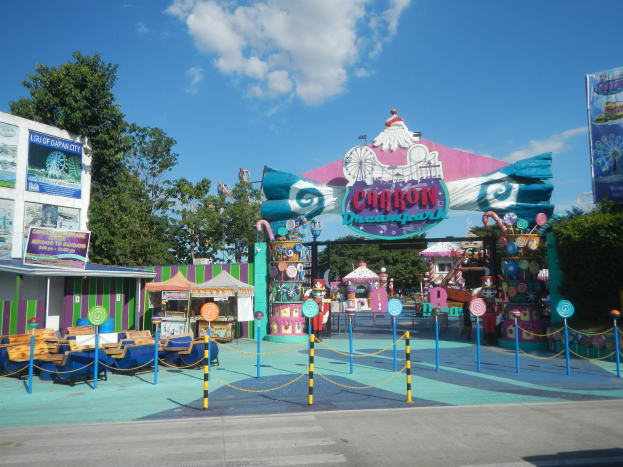 Karussell am Parkeingang mit einer Straße darunter, eingezäunter Bereich, Buden, Banner, Bäume und bewölkter Himmel.