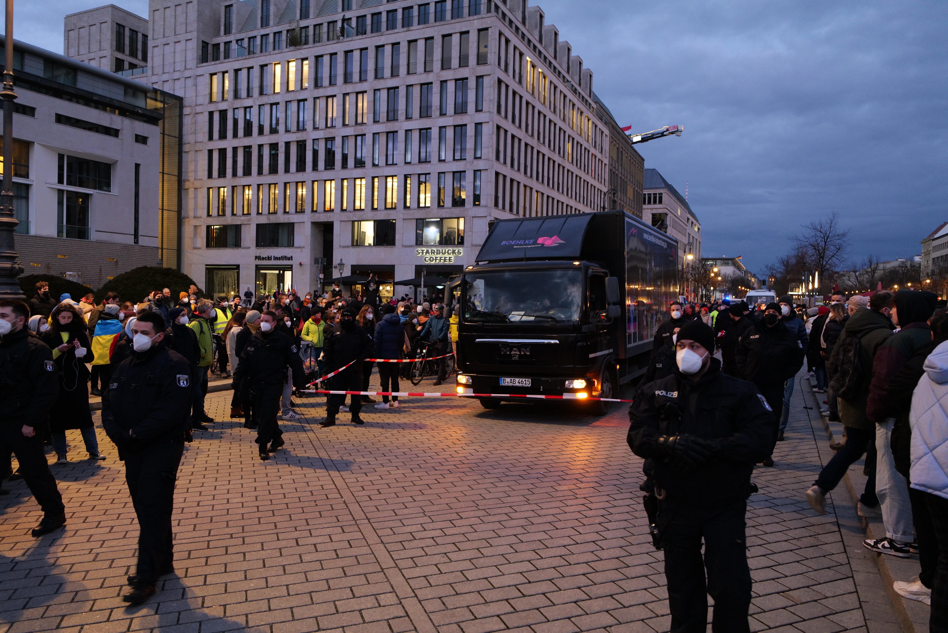 Eine Gruppe von Menschen steht in der Nähe eines Lastwagens auf einer Straße, die von Gebäuden, Laternenmasten, Bäumen und einem bewölkten Himmel gesäumt ist, wobei einige eine Mütze und eine Maske tragen und ein Band auf einem Pfahl im Vordergrund zu sehen ist.