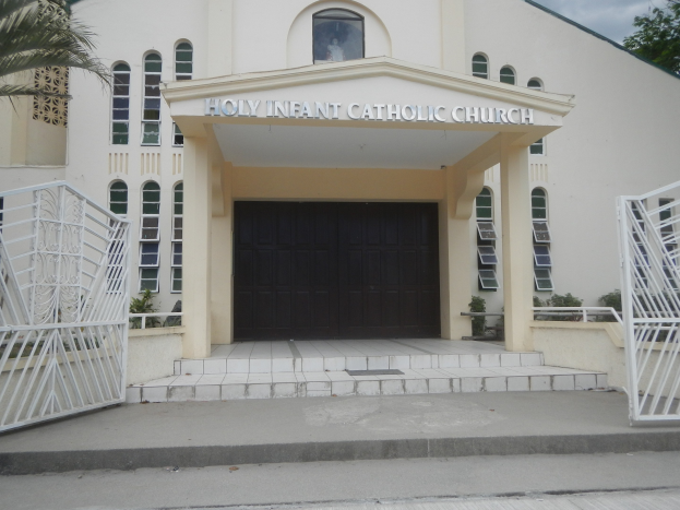 Äußenansicht der Heiligen-Kind-Katholischen Kirche mit Treppe, Geländern, umgebender Vegetation und einem bewölktem Himmel.