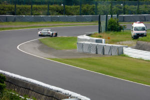Ein Rennwagen fährt eine Rennstrecke entlang mit einem Polizeiwagen im Hintergrund, umgeben von Grünflächen und Absperrungen, mit Text am unteren Bildrand.
