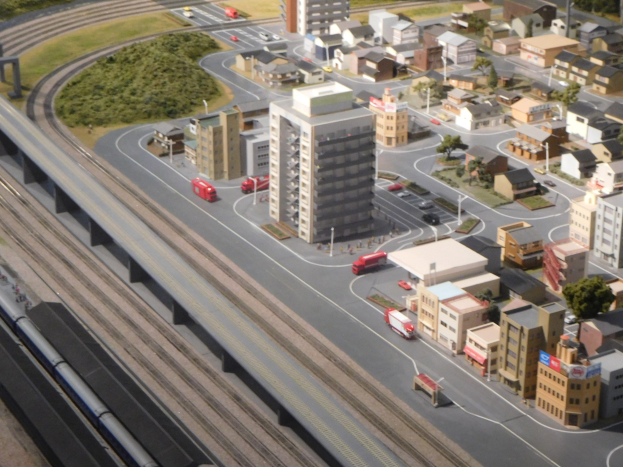 Ein Miniaturstadtmodell mit einem Zug auf Schienen, umgeben von Gebäuden, Bäumen, Straßen, Fahrzeugen, Strommasten und Gras, mit einem Wolkenkratzer im Hintergrund.