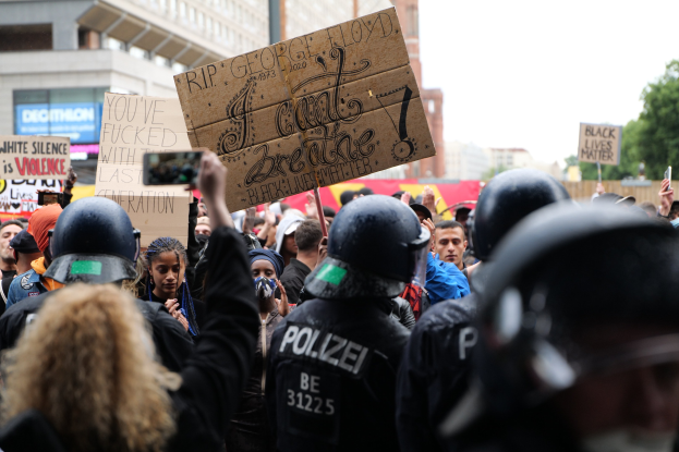 Eine Gruppe von Menschen mit Helmen und Schildern und Handys während einer Demonstration vor Gebäuden und Bäumen.