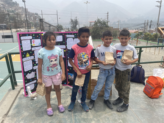 Gruppe von Kindern auf einem Basketballfeld, die Papiertüten halten, mit Vorräten auf dem Boden und einer Tafel mit Text links, vor einem Hintergrund aus Zäunen, Pfosten, Straßenlaternen, Bäumen, Gebäuden, Hügeln und dem Himmel.