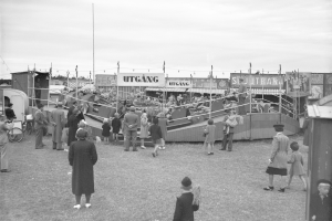 Ein Schwarz-Weiß-Foto einer Menschenmenge auf einem Fasching mit Buden, Pfählen und Bäumen im Hintergrund unter einem klaren Himmel.