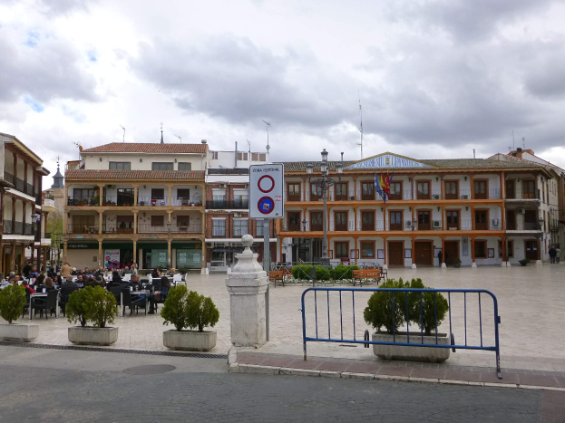 Ein belebter Stadtplatz mit Menschen auf Stühlen und Stehenden, Topfpflanzen, einer Metallabsperrung, einem Schild an einem Pfahl, Straßenlaternen mit Flaggen, umliegenden Gebäuden und einem bewölkten Himmel.