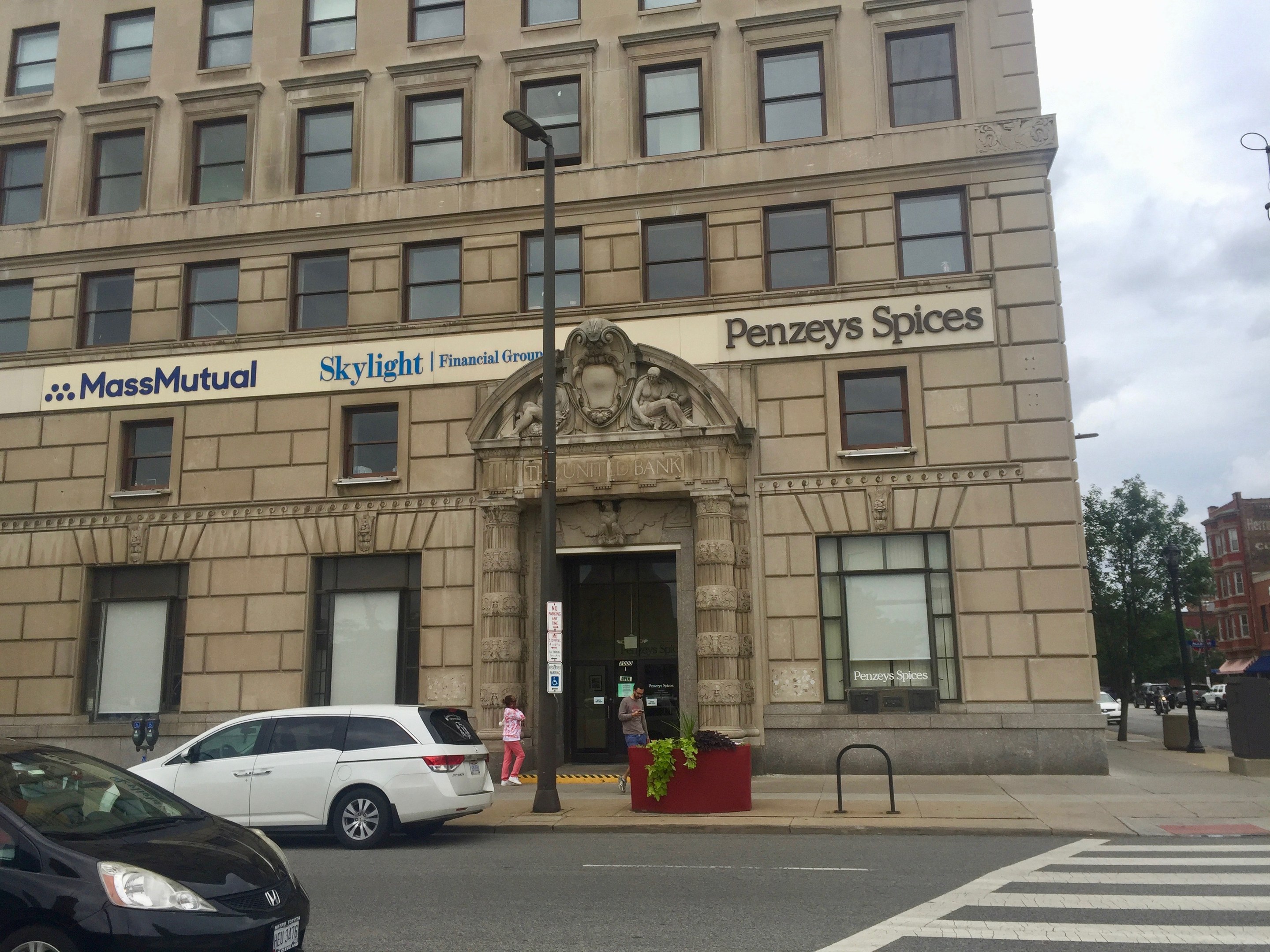 Großes Gebäude mit dem Massachusetts Mutual Financial Group-Logo, parkende Autos davor, Straßenlaternen, Lichter, fahrende Fahrzeuge, Fußgänger auf dem Gehweg, Grünpflanzen und ein bewölkter Himmel.