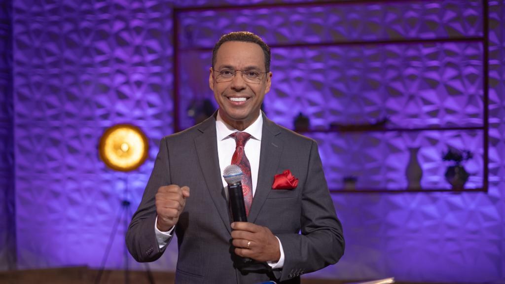 Stephen Colbert, Moderator von "The Daily Show with Trevor Noah", steht auf einer Bühne und hält ein Mikrofon in der Hand, trägt einen Anzug, Krawatte und eine Brille, während im Hintergrund ein Scheinwerfer und ein Ständer zu sehen sind.