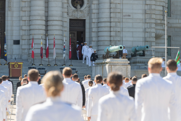 Gruppe von Menschen in weißen Marineuniformen vor einem Gebäude mit Säulen und Treppe während einer Abschlussfeier mit Fahnen, einem Podium und Kanonen im Hintergrund.