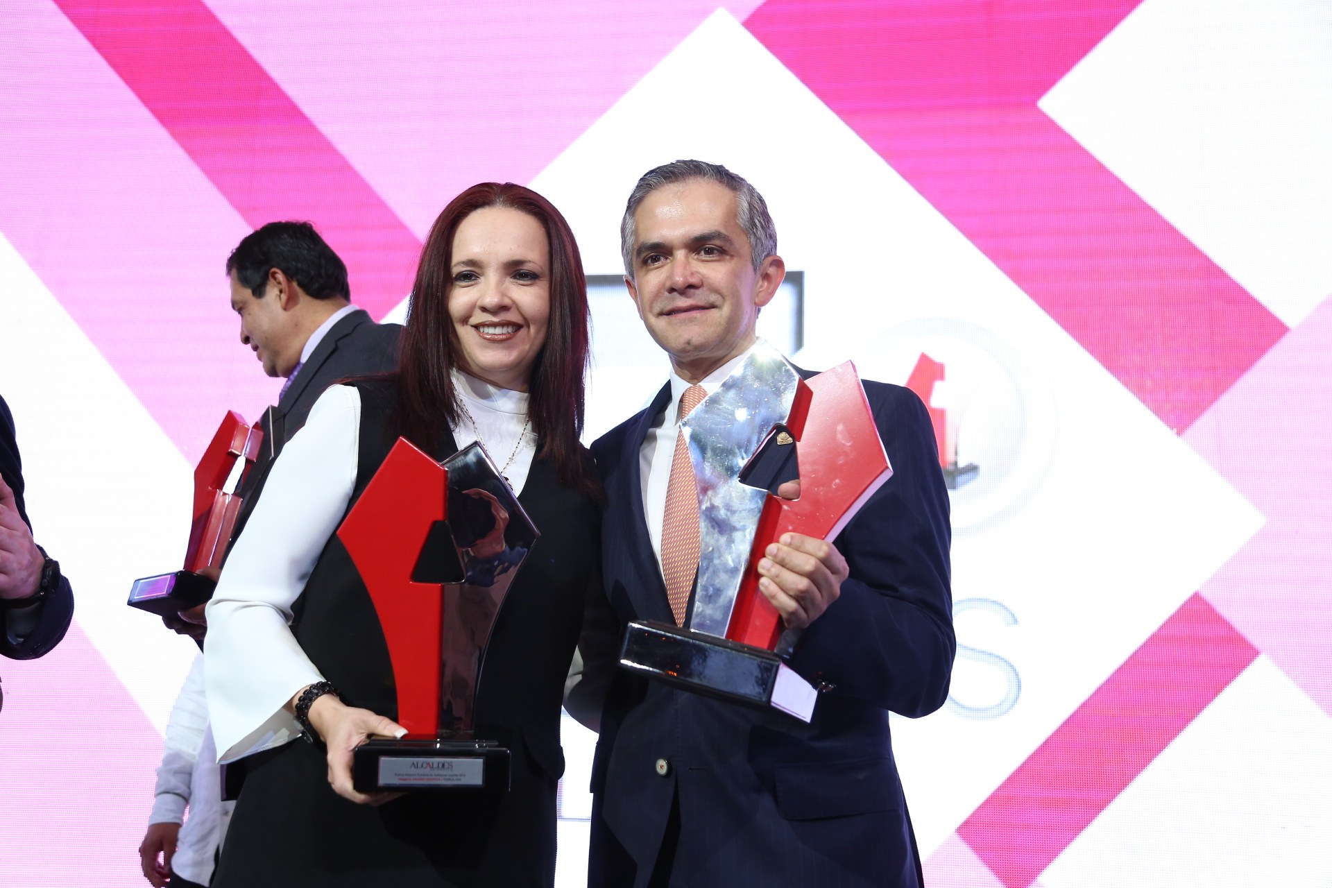 Zwei Personen auf einer Bühne mit Trophäen in den Händen, die lächeln, mit einem Bildschirm im Hintergrund.