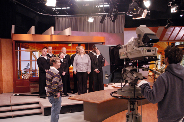 Eine Gruppe von Menschen in einem Fernsehstudio, wobei eine Person eine Kamera hält und andere in der Nähe eines beleuchteten Fernsehgeräts stehen.