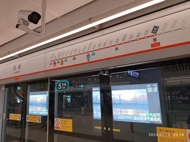 Ein Zug an einem Bahnhof mit einer Überwachungskamera an der Decke, umgeben von Glastüren, Texttafeln und anderen Gegenständen, mit ein paar Menschen im Zug sichtbar.