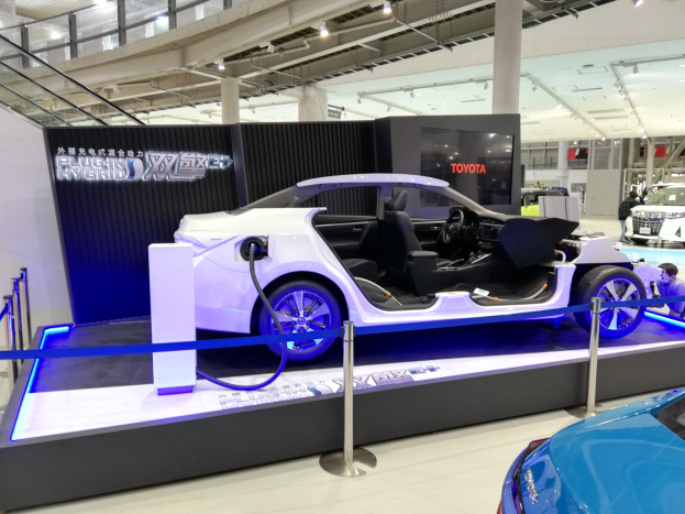 Ein silberner Toyota FT-1 Elektrokonzeptwagen auf der Tokyo Motor Show, umgeben von Absperrpollern mit Bändern, Informationsschildern, Deckenleuchten und anderen Fahrzeugen, mit Menschen im Hintergrund.