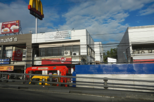 Ein McDonald's-Restaurant an einer Straßenecke, umgeben von Gebäuden, Werbetafeln, Strommasten, Fahrzeugen, Geländern, Bäumen und einem bewölkten Himmel.