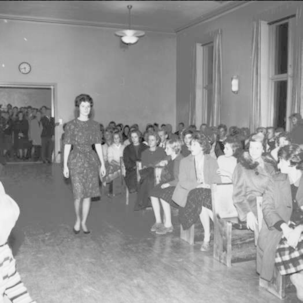 Eine Frau in einem schwarzen und weißen Kleid läuft über einen Laufsteg während einer Modenschau für Frauenwahlrecht, mit einer Menge, die auf Stühlen sitzt und einer Uhr und Lampen an den umliegenden Wänden.