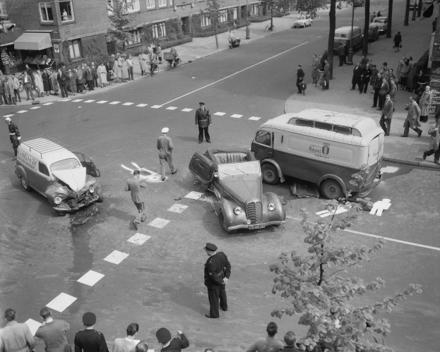 Schwarz-weiß-Szene eines Autounfalls an einer Straßenkreuzung mit einem Van, mehreren Fahrzeugen und einer Gruppe von Schaulustigen, vor Gebäuden, Bäumen, Pfählen und Texttafeln.