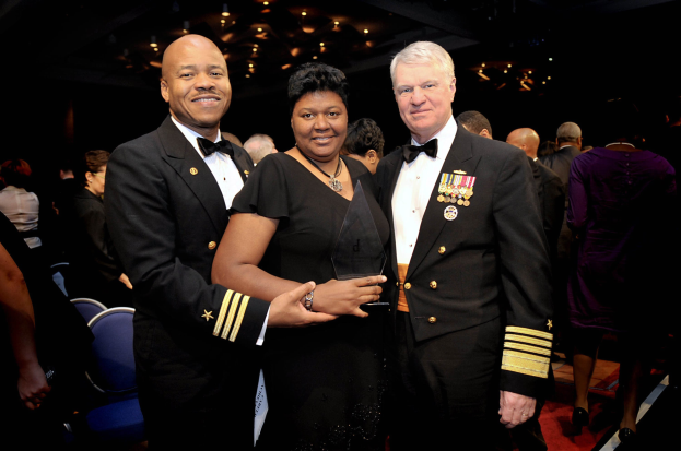 Zwei Männer in schwarzen Blazern und weißen Hemden stehen neben einer Frau in einem schwarzen Kleid, die eine Trophäe auf einem roten Teppich hält, während andere im Hintergrund sitzen und stehen unter beleuchteter Decke.