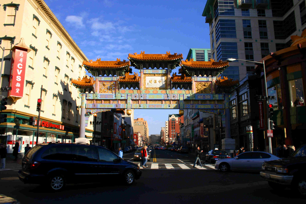 Eine Straße mit fahrenden Fahrzeugen, Menschen, die an einer Ampel Über die Straße gehen, ein Tor im Hintergrund, Gebäude auf beiden Seiten und eine Ampel unter einem klaren Himmel.