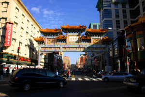 Eine Straße mit fahrenden Fahrzeugen, Menschen, die an einer Ampel Über die Straße gehen, ein Tor im Hintergrund, Gebäude auf beiden Seiten und eine Ampel unter einem klaren Himmel.