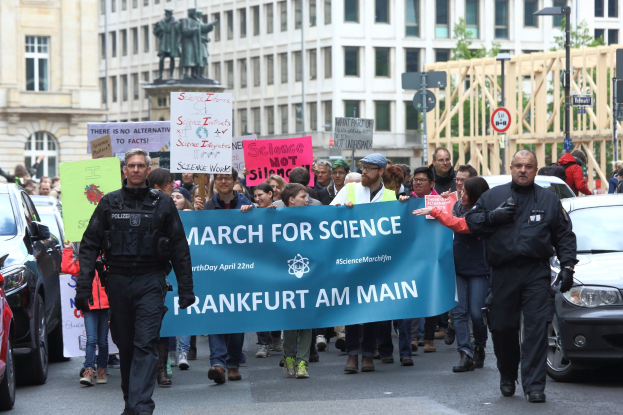 Gruppe von Menschen marschiert mit einem "Science March Frankfurt am Main"-Schild die Straße entlang, Autos und Gebäude im Hintergrund.