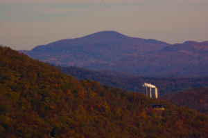 Eine Fabrik mit Rauch aus dem Schornstein vor einem Hintergrund von Bergen mit Bäumen unter einem bewölkten Himmel.
