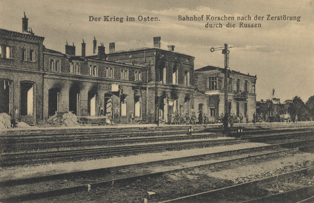 Schwarzes und weißes Foto des Bahnhofs Korschen in Ostern, Deutschland, das Schienenstränge, Bahnhofsgebäude, Menschen, Bäume und einen Himmelhintergrund mit der Aufschrift "Bahnhof Korschen in Ostern, Deutschland" oben und unten zeigt.
