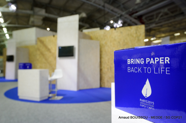 Messestände mit einem blauen und weißen Schild, auf dem "Bring Paper Back to Life" steht, auf der rechten Seite, im Hintergrund sind leicht unscharfe Stühle, Tische, ein Wandfernseher und Deckenleuchten zu sehen.