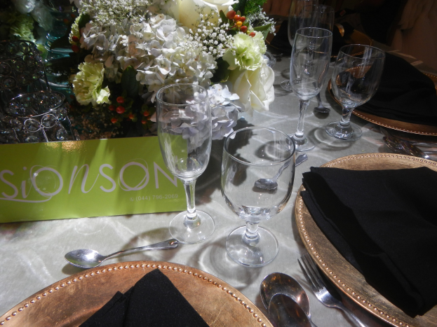 Ein formell gedeckter Esstisch mit goldenen Untersetzern, schwarzen Servietten, Besteck, Gläsern und einem Blumenstrauß, auf dem eine Tafel mit Text steht.