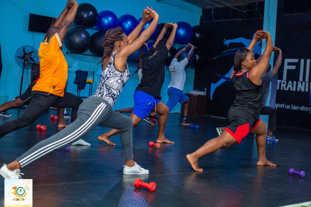 Gruppe von Menschen, die Yoga in einem Fitnessstudio machen, mit Hanteln auf dem Boden, einem Banner mit der Aufschrift 'Fitness-Training in Ghana' im Hintergrund und diversen Gegenständen wie Luftballons und einem Ventilator.