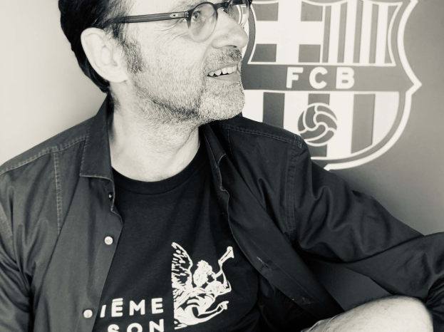 Ein Mann mit Brille und schwarzem Hemd lächelt, während er vor einem schwarz-weißen FC Barcelona-Logo sitzt.
