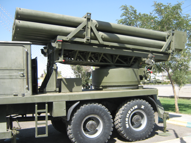 Ein militärisches Lastauto mit einer Rakete fährt eine Straße entlang, umgeben von Gras, Bäumen, Gebäuden und einem Maschendrahtzaun.