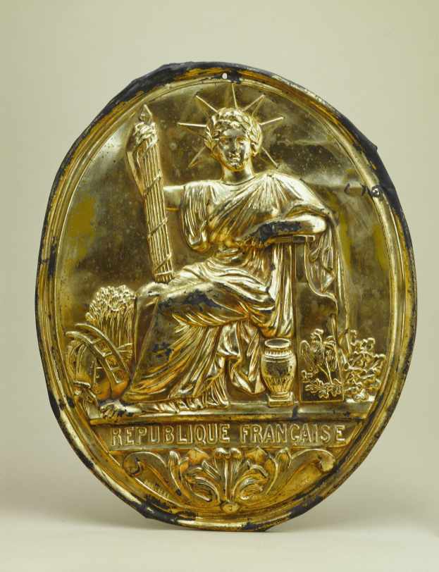 Goldener kreisförmiger Schild mit einer Miniaturstatue der Freiheitsstatue und eingraviertem Text auf weißem Hintergrund.