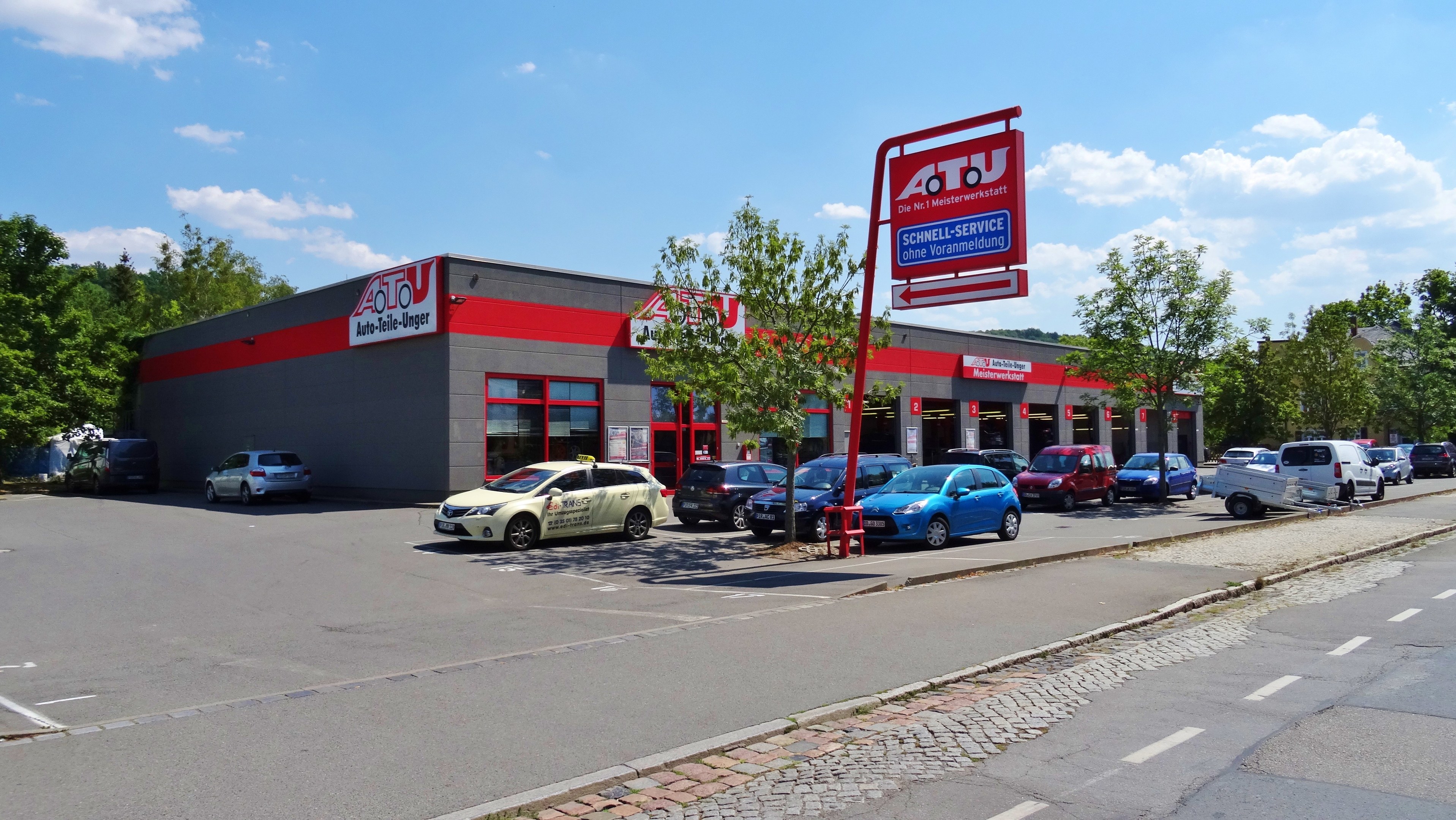 Autohaus mit parkenden Autos, umgeben von Gebäuden, Bäumen, Strommasten, Schildern und einer Straße unter einem bewölkten Himmel.
